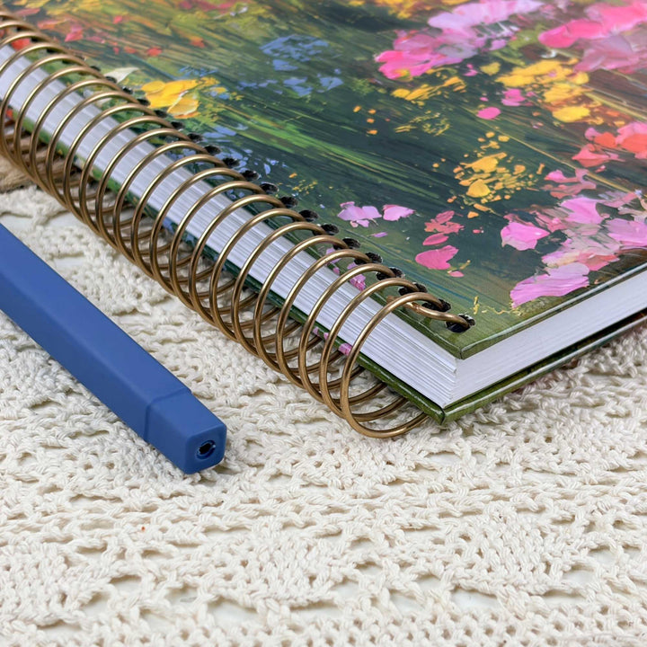 A spiral-bound notebook with a colorful floral cover and a gold wire coil, placed on a cream lace doily next to a blue pen.