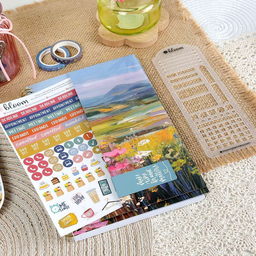 A Bloom brand planner sticker sheet featuring a colorful landscape illustration and various decorative stickers, styled on a beige woven mat alongside a clear plastic grid template and other stationery accessories.