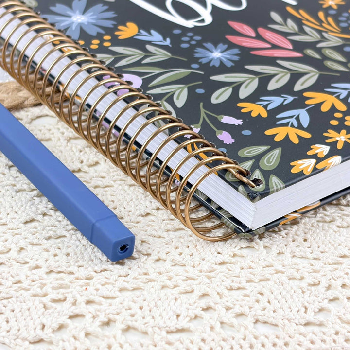 A close-up of a spiral-bound notebook with a floral-patterned cover and a gold wire spiral, placed on a cream crocheted surface next to a blue pen.
