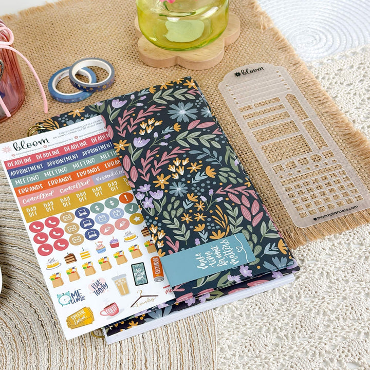 A floral planner sticker sheet and matching planner inserts are arranged on a woven beige surface with a clear plastic divider sheet nearby. The sticker sheet has many colorful stickers and labels, and the planner insert has a dark navy background with pink, green, yellow, and purple floral patterns.