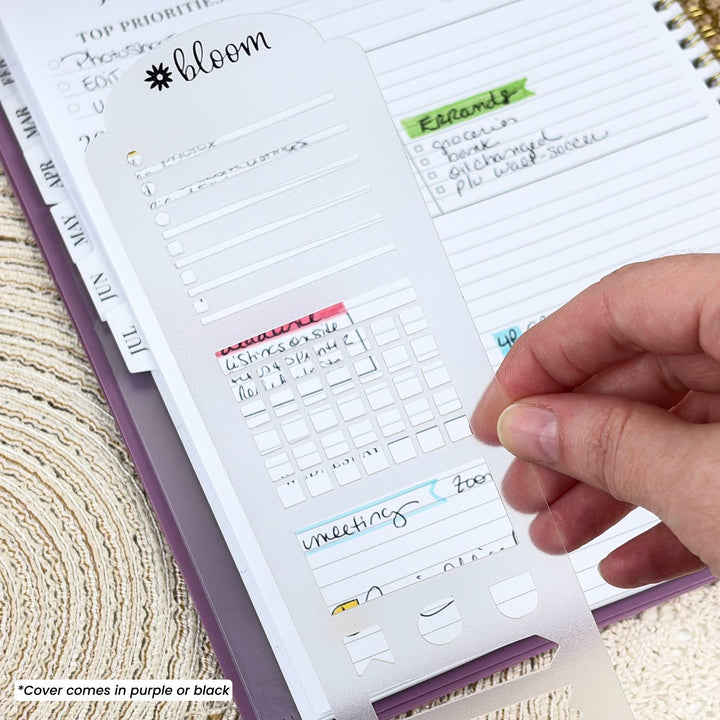 A close-up of a hand holding a white planner sticker sheet with checklist and calendar planner inserts, placed over an open purple planner. The sheet has the brand name 'bloom' printed at the top, and a small text note in the corner says '*Cover comes in purple or black'.