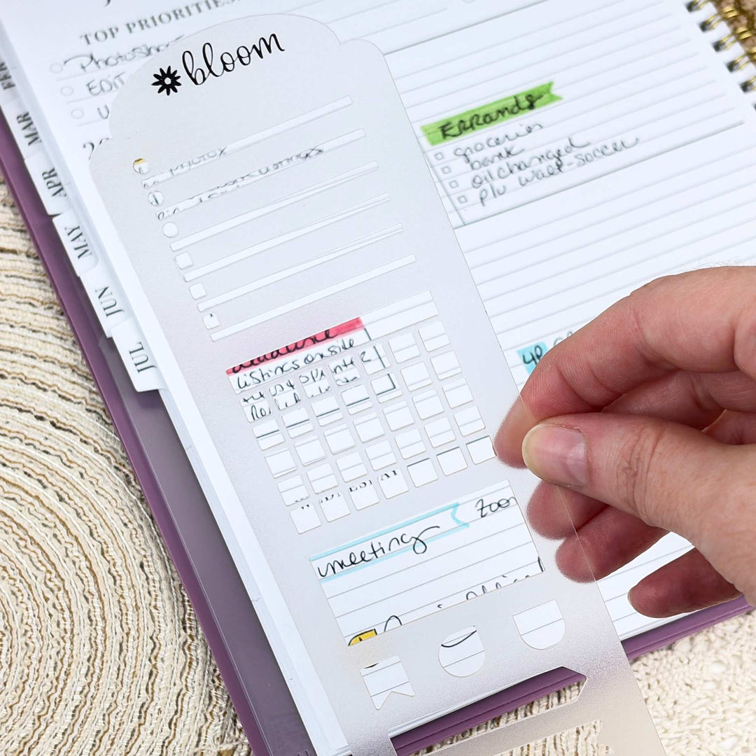 A hand holds a clear plastic planner page divider with the brand name 'bloom' and pre-cut slots, positioned over an open purple planner.