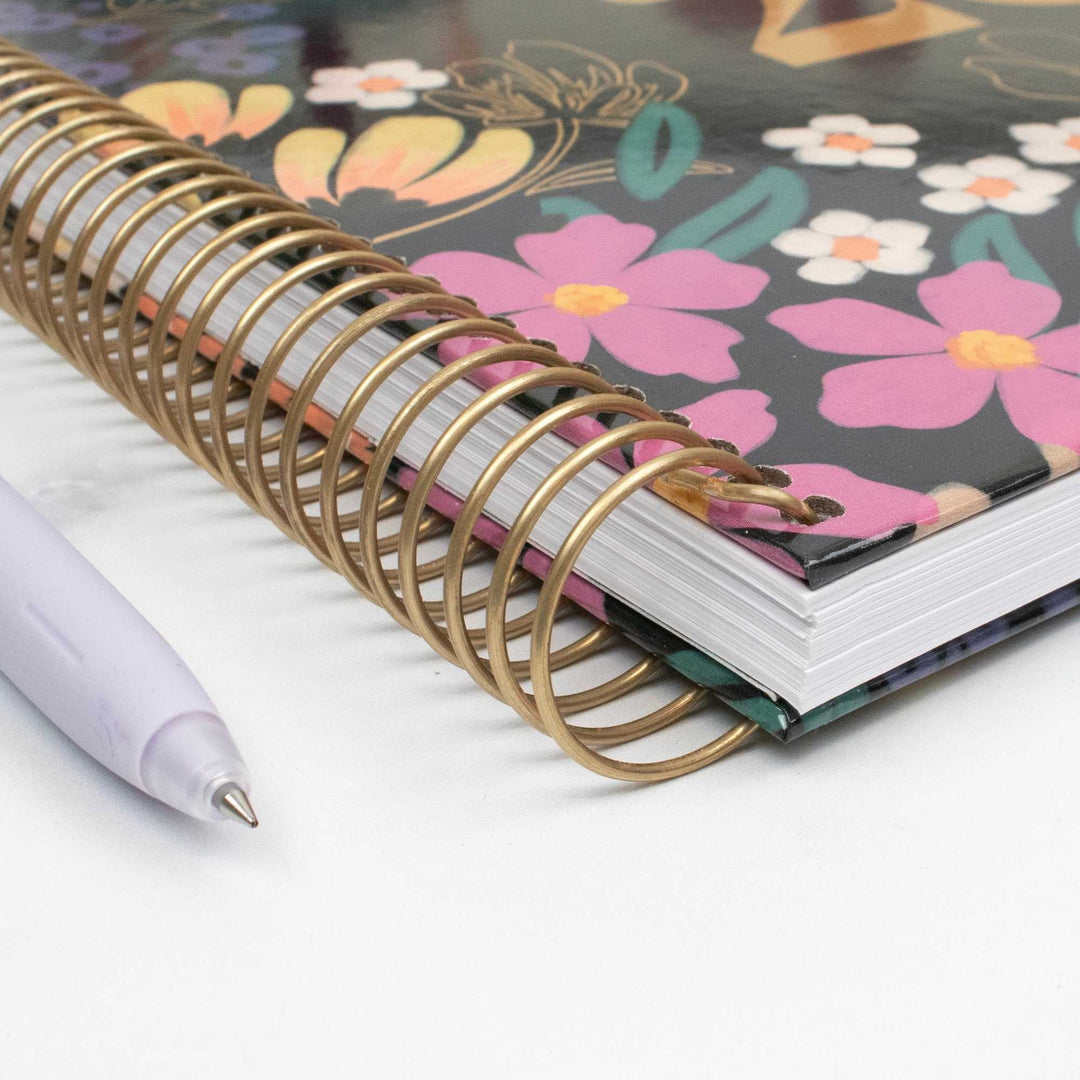 A close-up of a spiral-bound notebook with a metallic gold wire coil and a floral-patterned cover featuring pink, yellow, and white flowers on a dark background. A light lavender pen rests on the white surface beside the notebook.