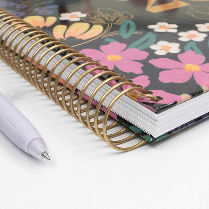 A close-up of a spiral-bound notebook with a metallic gold wire coil and a floral-patterned cover featuring pink, yellow, and white flowers on a dark background. A light lavender pen rests on the white surface beside the notebook.
