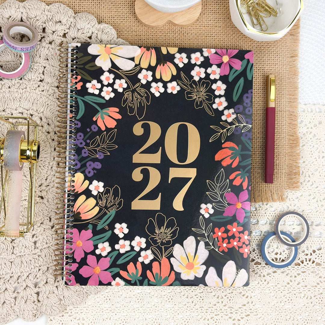 A spiral-bound 2027 planner with a black cover decorated with colorful floral illustrations and large gold '2027' text, styled on a lace mat with stationery props.