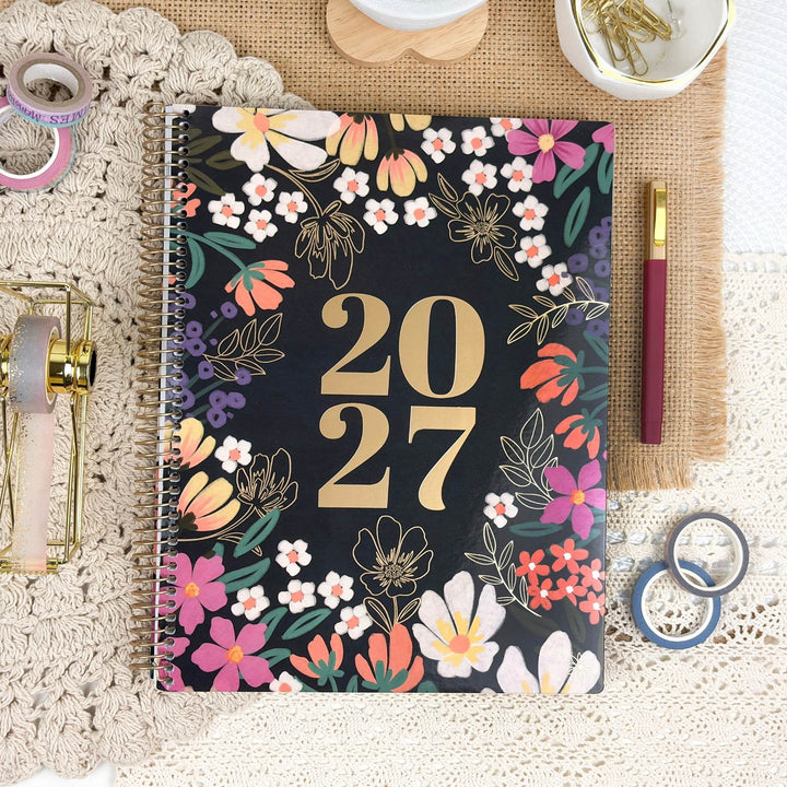 A spiral-bound 2027 planner with a black cover decorated with colorful floral illustrations and large gold '2027' text, styled on a lace mat with stationery props.