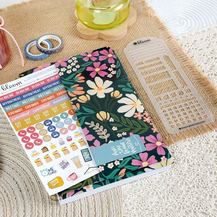 A floral planner cover or notebook with a sticker sheet and a clear plastic planner divider placed on top, styled on a woven beige mat with a glass of green drink in the background.