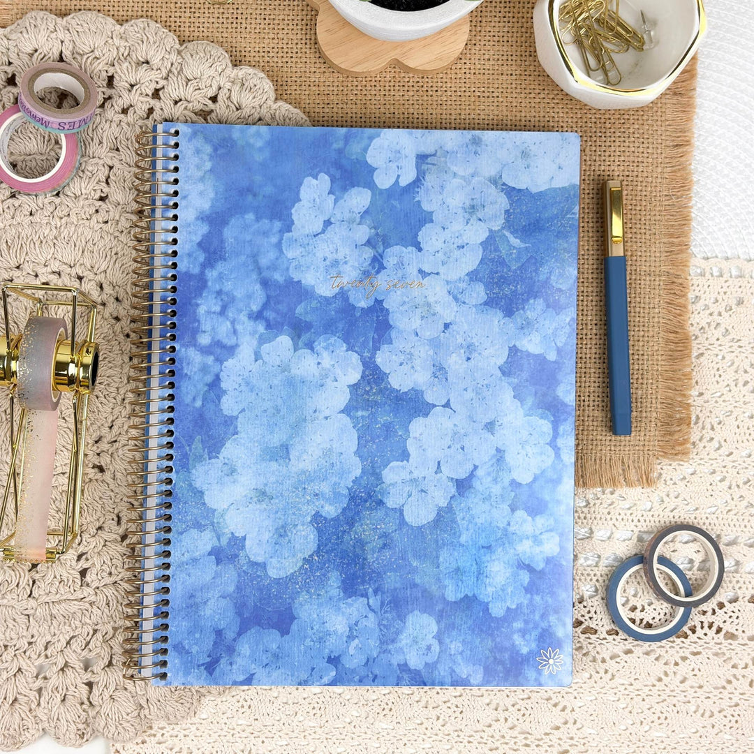 A spiral-bound notebook with a blue watercolor floral cover lies on a beige lace surface, surrounded by stationery props including a gold pen, washi tapes, paper clips, and a small cup.