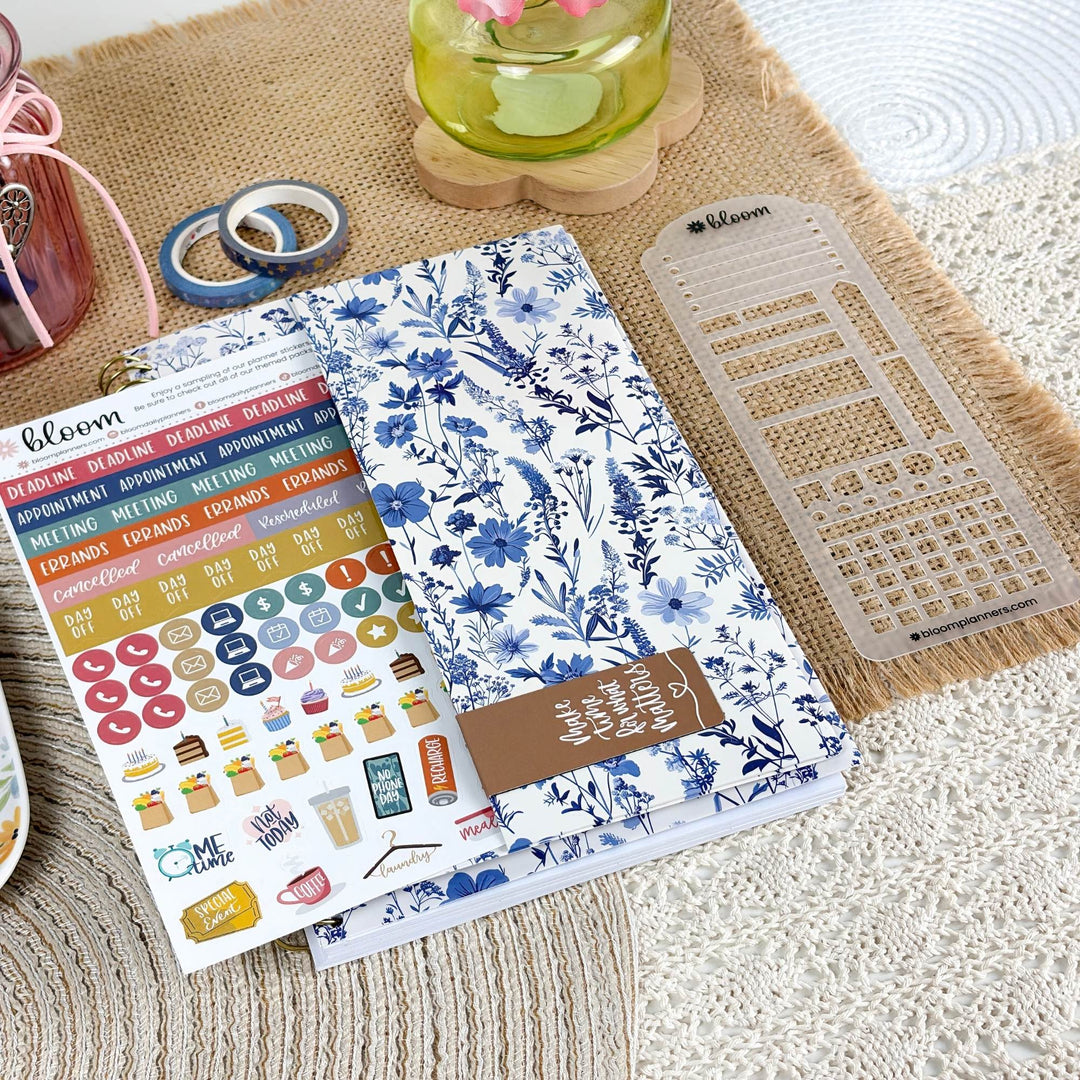 A planner sticker sheet and matching floral planner cover are arranged on a burlap mat with planner accessories around them. The cover has a blue and white floral print, and the sticker sheet includes colorful daily headers, checklist stickers, and small illustrated icons.