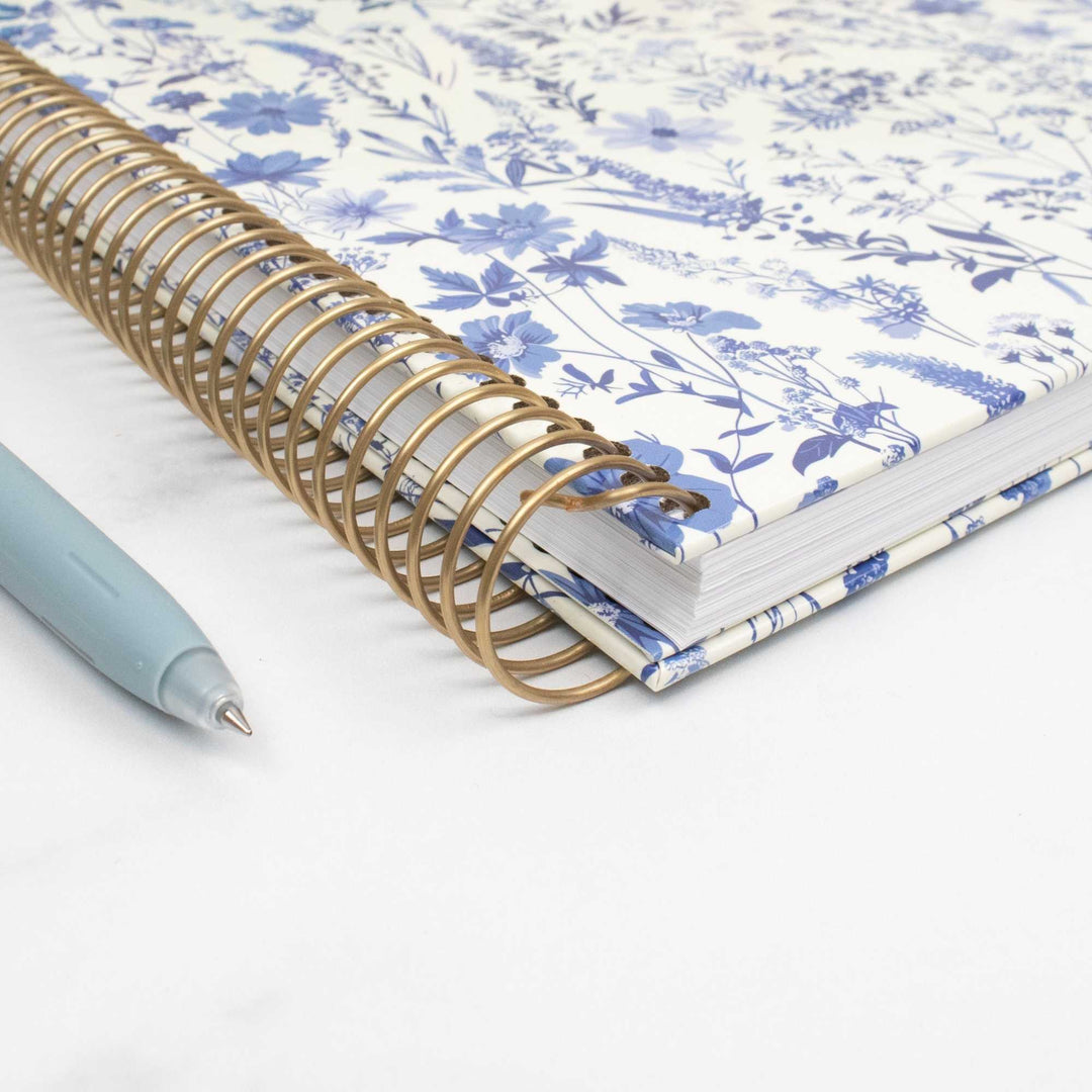 A spiral-bound notebook with a blue and white floral patterned cover, shown with a gold-tone spiral binding and a light blue pen on a white surface.