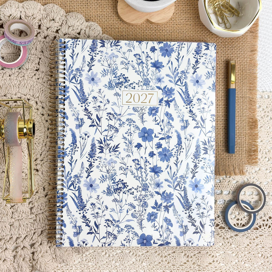 A 2027 spiral-bound planner with a blue and white floral pattern, styled in a flat-lay arrangement with stationery accessories on a beige lace background.