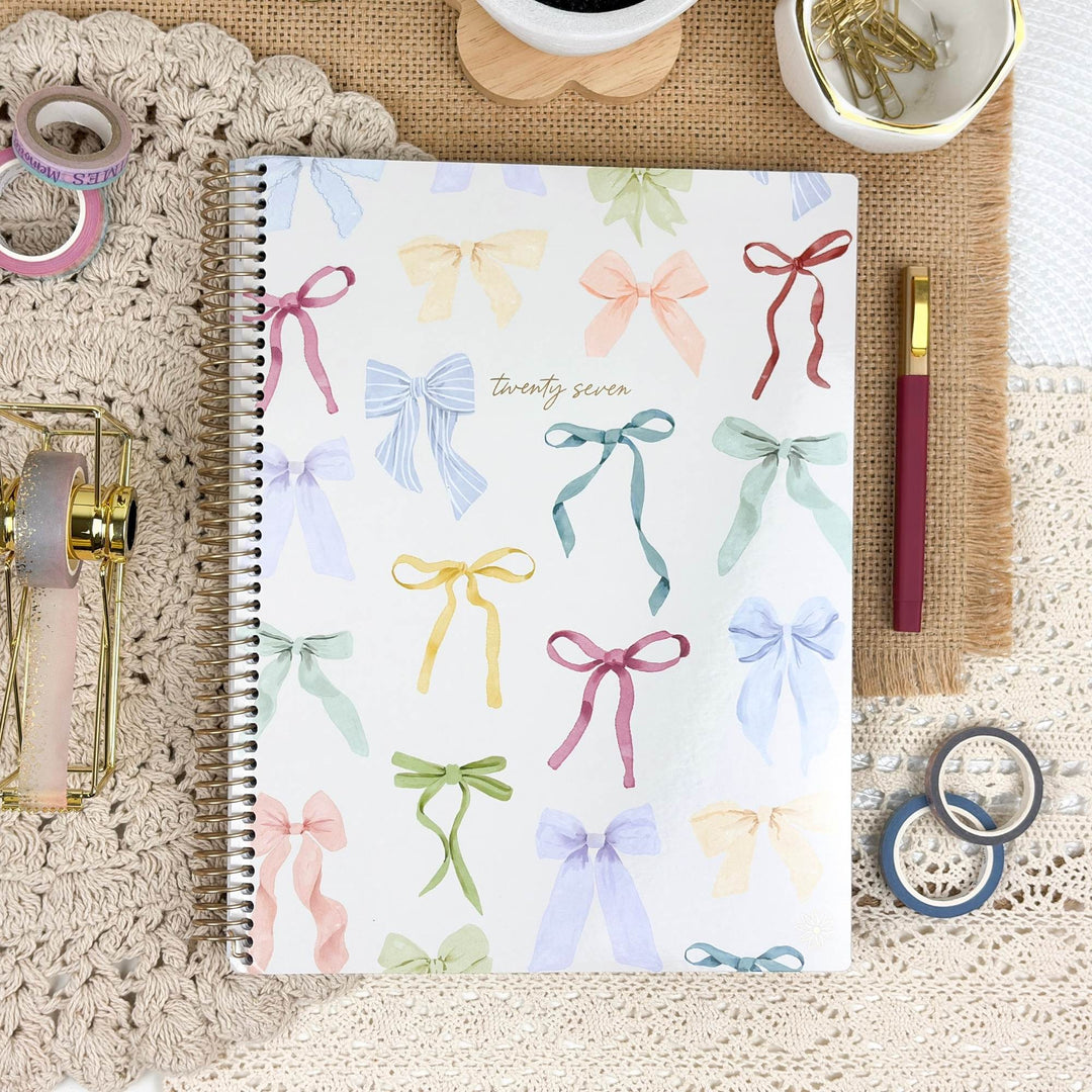 A spiral-bound planner or notebook with a white cover decorated with colorful watercolor ribbon bows, styled in a flat lay on a lace surface with pens, washi tape, rings, and other stationery props.