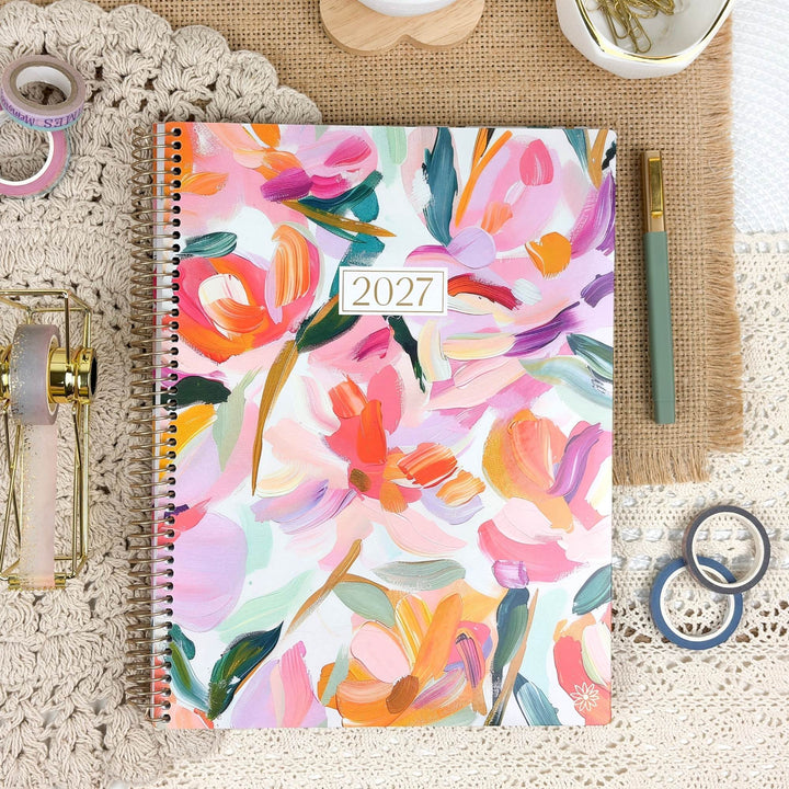 A 2027 spiral-bound planner with a colorful floral watercolor cover, styled in a flat-lay arrangement on a lace tablecloth with a burlap mat, pen, washi tape, and small wooden accessories.