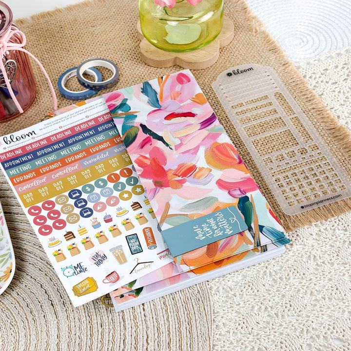 A styled flat-lay of a planner sticker sheet and matching planner accessories on a woven beige mat. The main product is a floral planner sticker sheet with pink, orange, blue, and green watercolor flowers, alongside a matching floral planner cover, a clear plastic grid divider, and washi tape.