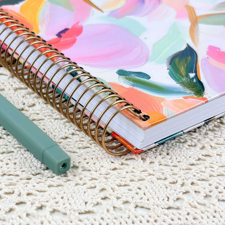 A spiral-bound notebook with a colorful abstract floral cover lies on a cream lace doily, with a mint green pen resting beside it.