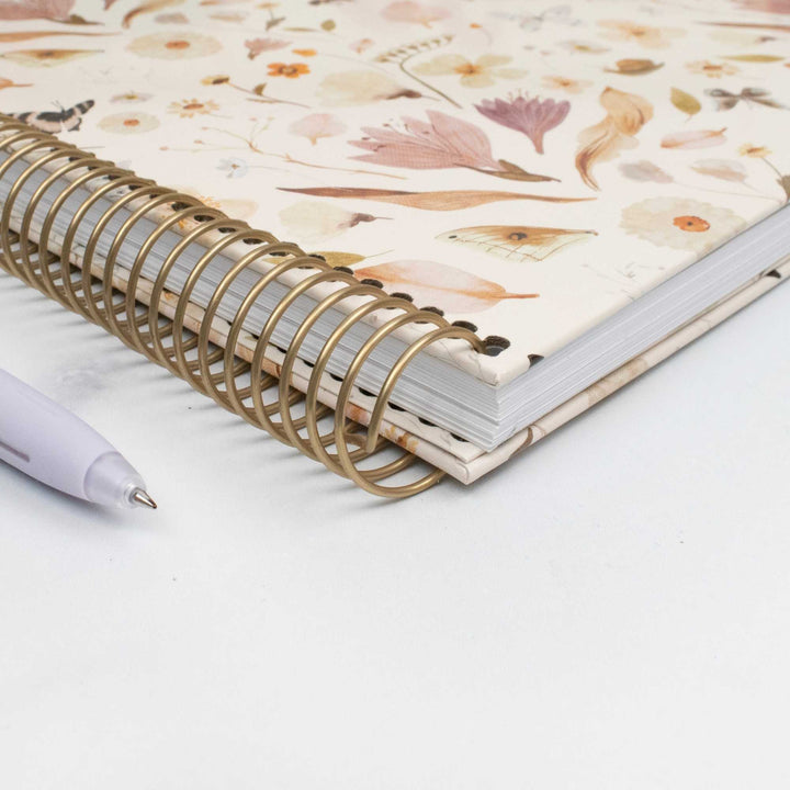 A close-up of a spiral-bound notebook with a cream floral cover and gold wire binding, placed on a white surface with a light lavender pen beside it.