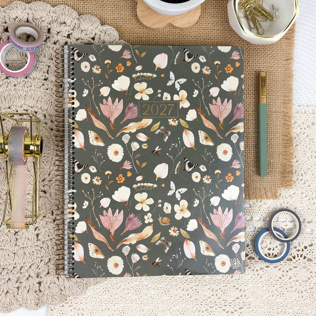 A 2027 spiral-bound planner with a dark gray floral cover, styled in a flat-lay arrangement with stationery props on a lace cloth and burlap mat.