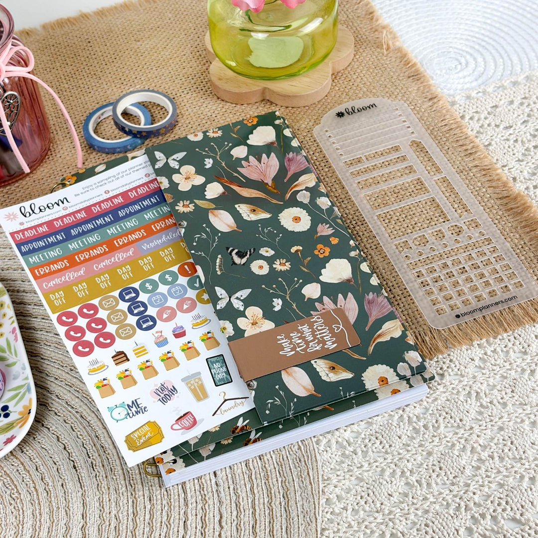 A floral planner sticker sheet and matching planner accessories are arranged on a woven mat. The main item is a dark green planner cover or notebook with white and multicolor floral print, a brown tag reading 'I love you very much', a clear plastic grid template, and a sheet of colorful stickers.
