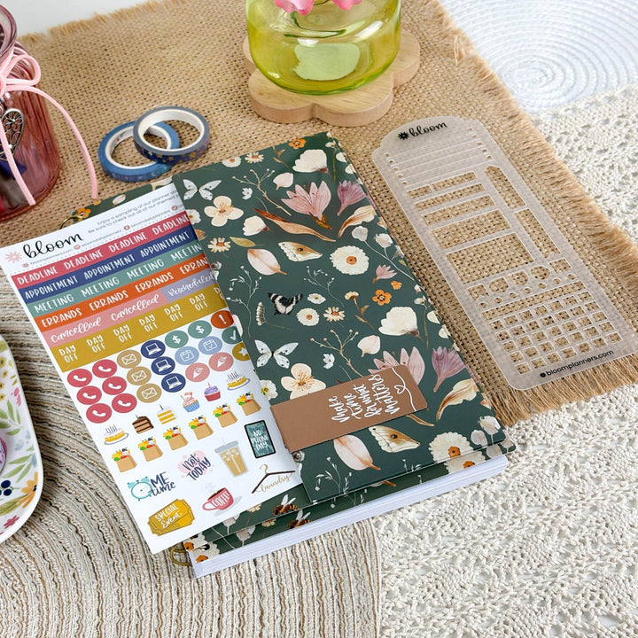 A floral planner sticker sheet and matching planner accessories are arranged on a woven mat. The main item is a dark green planner cover or notebook with white and multicolor floral print, a brown tag reading 'I love you very much', a clear plastic grid template, and a sheet of colorful stickers.
