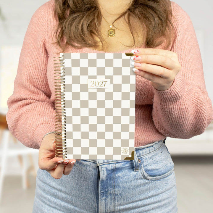 A person in a pink knit sweater holds a spiral-bound 2027 planner with a beige and white checkerboard cover in front of their torso.