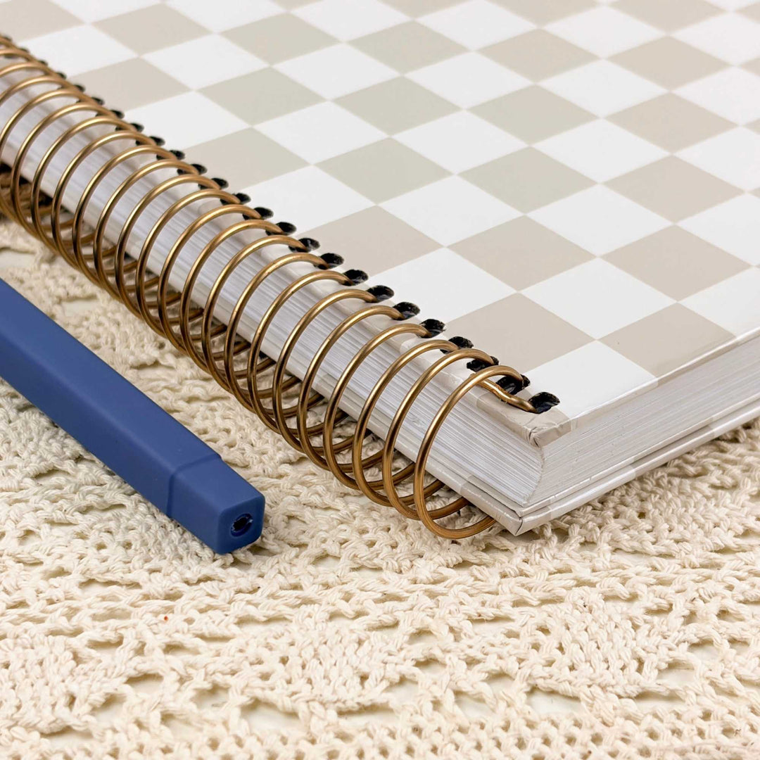 A spiral-bound notebook with a beige and white checkerboard cover, shown with a gold wire coil, resting on a cream-colored crocheted textile next to a blue pen.