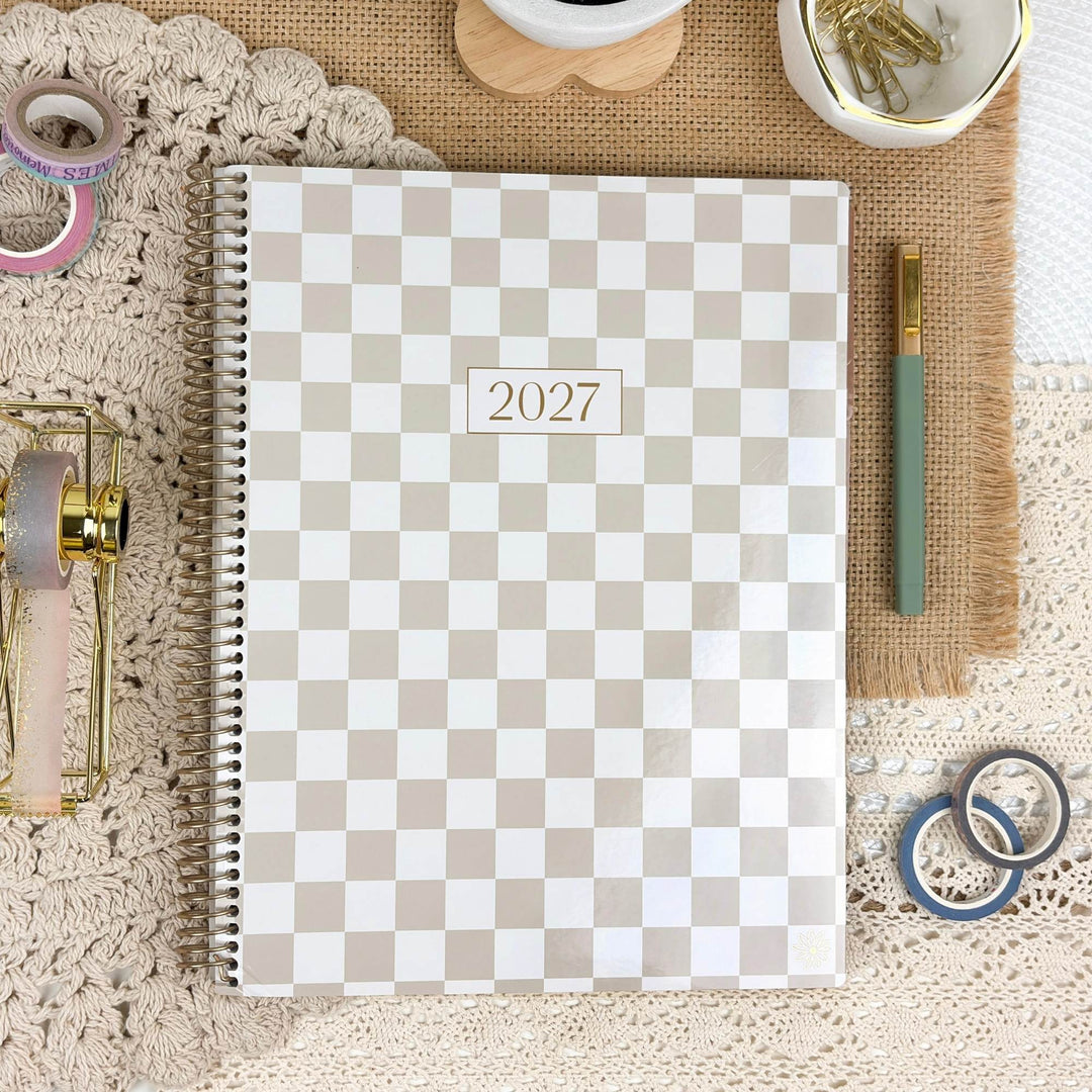 A spiral-bound 2027 planner with a beige and white checkerboard pattern, styled in a flat-lay arrangement on a lace cloth with stationery props.