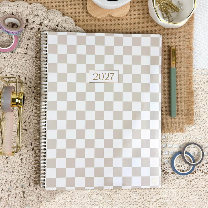 A spiral-bound 2027 planner with a beige and white checkerboard pattern, styled in a flat-lay arrangement on a lace cloth with stationery props.