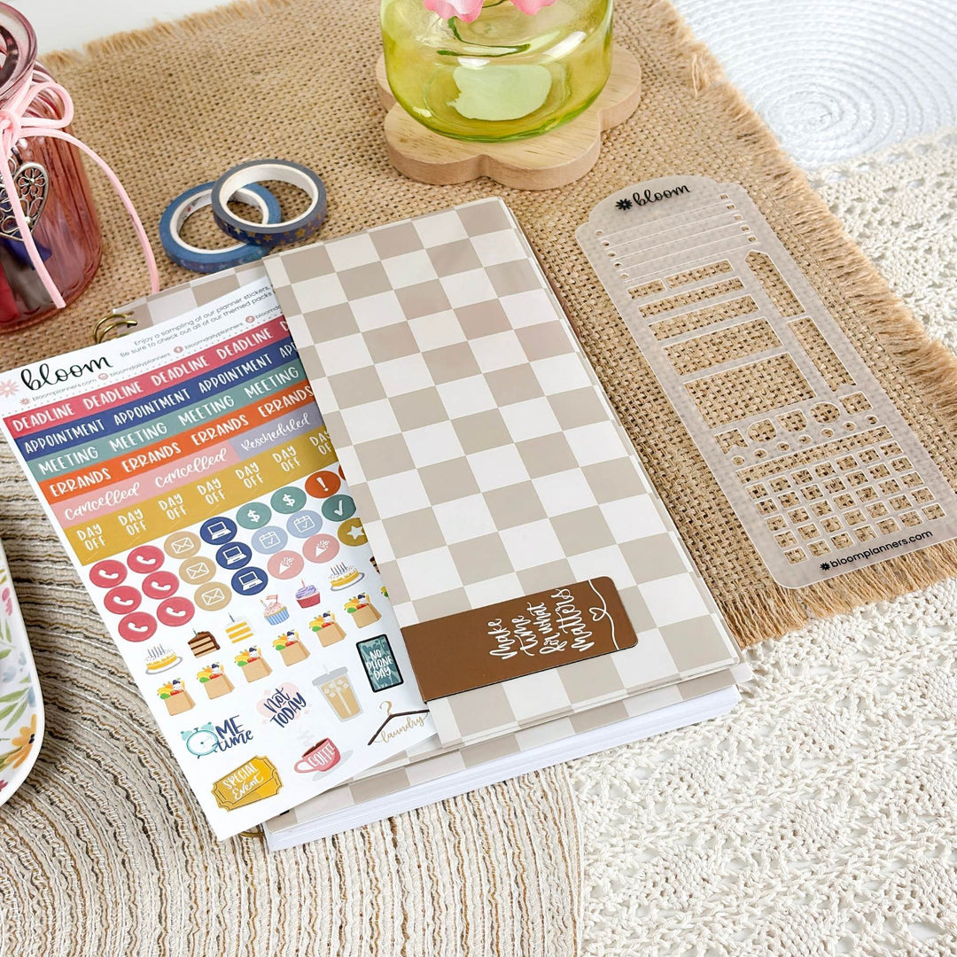 A planner sticker sheet and matching planner accessories arranged on a textured cream surface with a burlap mat. The main product appears to be a Bloom planner sticker set, with a checkered notebook cover, clear grid stickers, washi tape, and colorful sticker sheets visible.