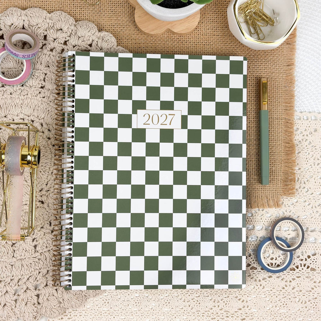 A spiral-bound 2027 planner with a green and white checkerboard cover, styled in a flat lay on a beige lace cloth with desk accessories including washi tape, a pen, gold paper clips, and a small plant.