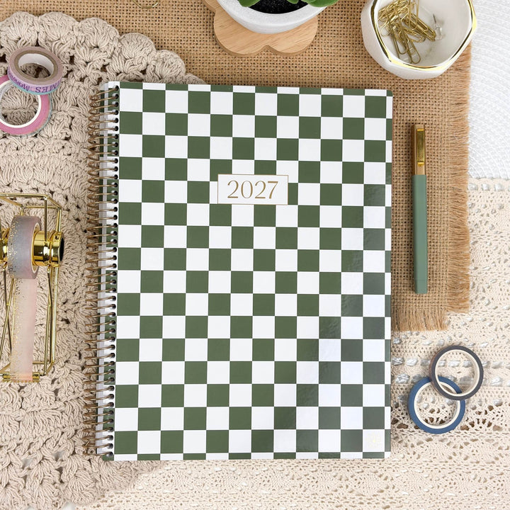 A spiral-bound 2027 planner with a green and white checkerboard cover, styled in a flat lay on a beige lace cloth with desk accessories including washi tape, a pen, gold paper clips, and a small plant.