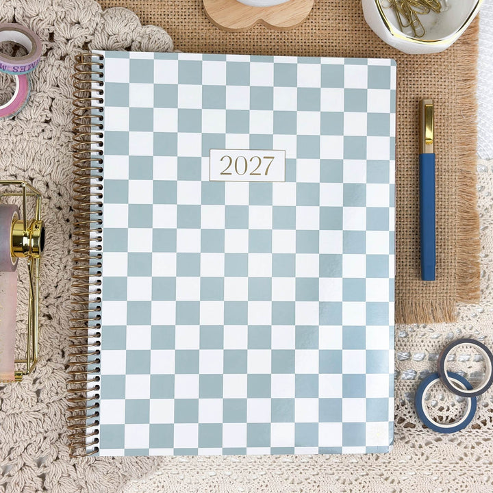 A spiral-bound 2027 planner with a light blue and white checkerboard pattern, styled in a flat-lay arrangement with a blue pen, washi tape, and other desk accessories on a textured beige background.