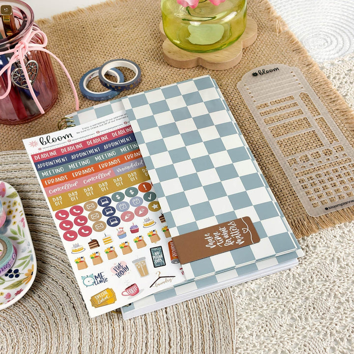 A planner sticker sheet set from the brand Bloom, featuring a light blue and white checkered cover, colorful daily headers, round icons, and cute illustrations like coffee cups and donuts, styled on a burlap mat with other desk accessories.