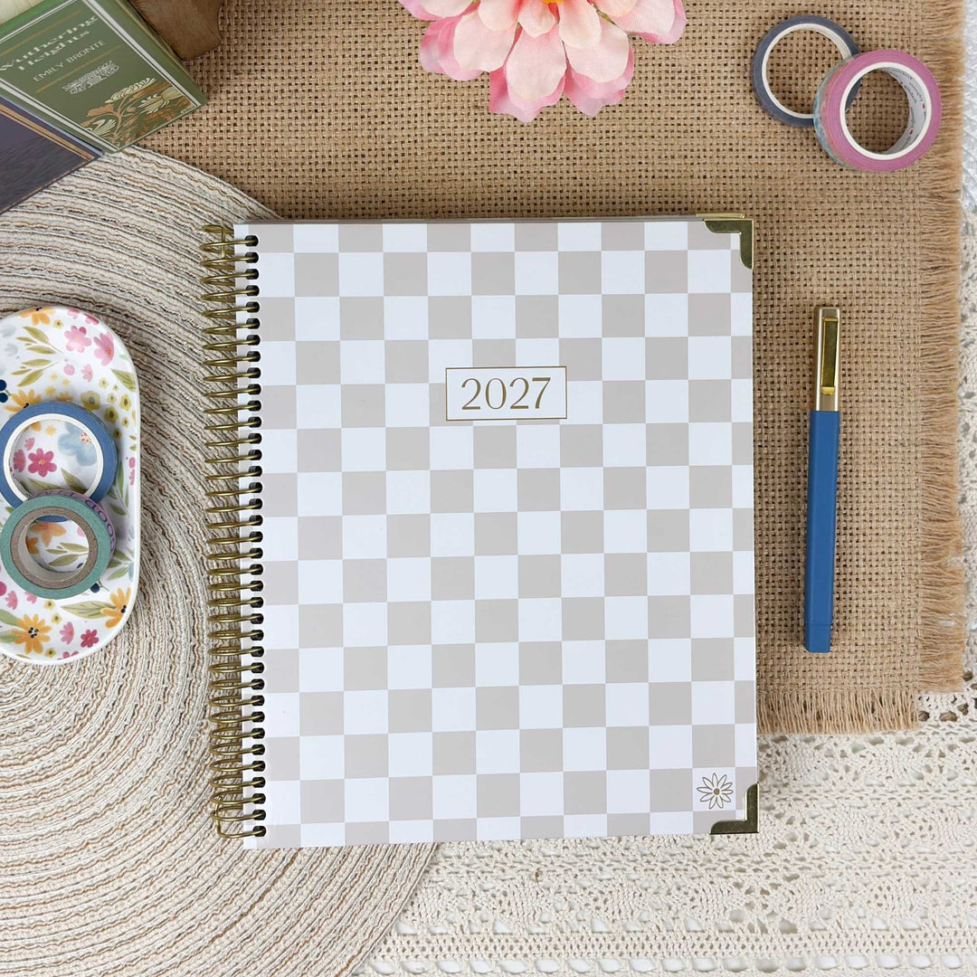 A spiral-bound 2027 planner with a beige and white checkerboard cover, styled in a flat-lay arrangement on a burlap mat with decorative washi tape, a pen, a flower, and a woven mat.