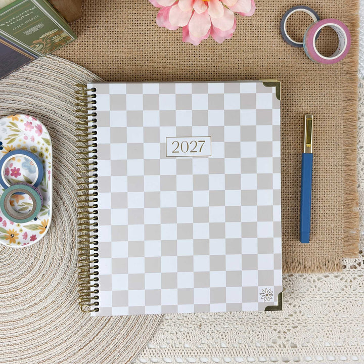 A spiral-bound 2027 planner with a beige and white checkerboard cover, styled in a flat-lay arrangement on a burlap mat with decorative washi tape, a pen, a flower, and a woven mat.