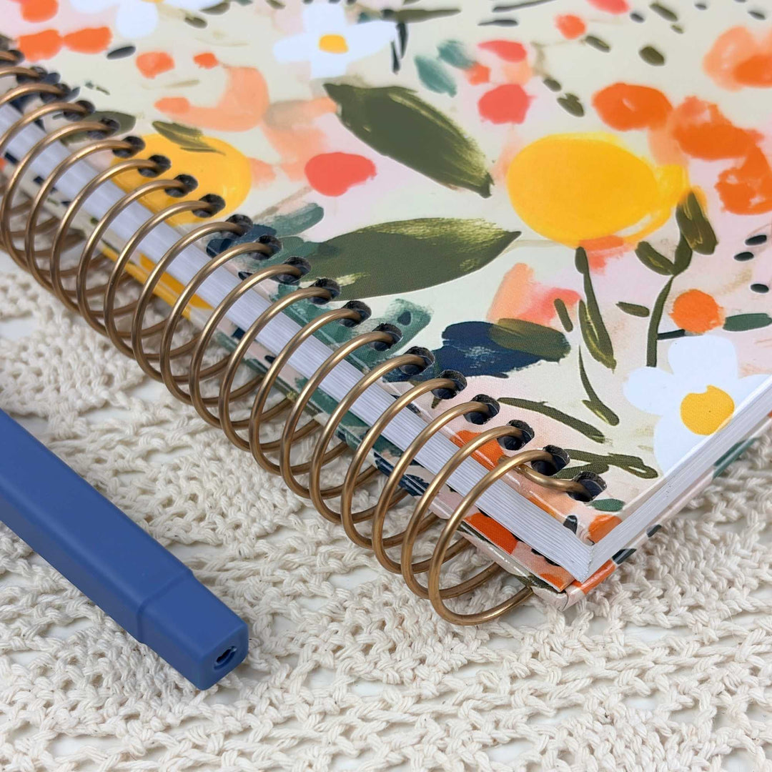 A close-up of a spiral-bound notebook with a colorful floral cover design, placed on a cream knit blanket with a blue pen nearby.