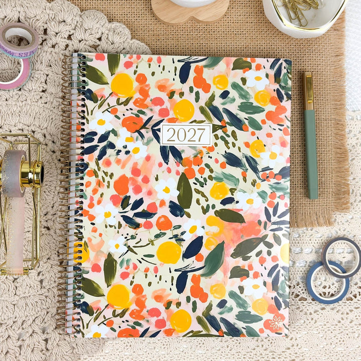 A spiral-bound 2027 planner with a colorful floral and citrus watercolor cover design, styled in a cozy desk flat-lay with a green pen, washi tape, and a small dish.