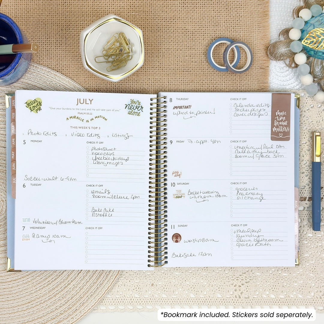 An open spiral-bound planner laid flat on a beige fabric surface, surrounded by small styled props including a blue cup, a dish with greenery, two rings, a beaded bracelet, and a pen. The planner pages are white with handwritten notes and printed weekly planner layouts.