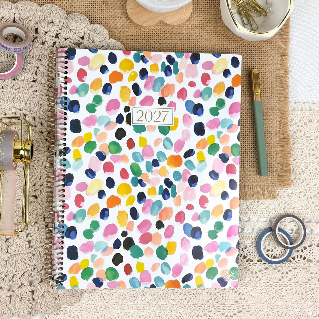 A 2027 spiral-bound planner with a colorful watercolor polka-dot pattern on a white cover, styled in a flat-lay arrangement with washi tape, a green pen, and decorative accessories on a burlap and lace background.