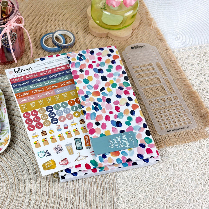 A styled flat-lay of a planner sticker kit from the brand bloom, featuring a white sticker sheet with colorful watercolor dots, a matching sticker book, and a clear plastic grid template on a burlap and lace background.