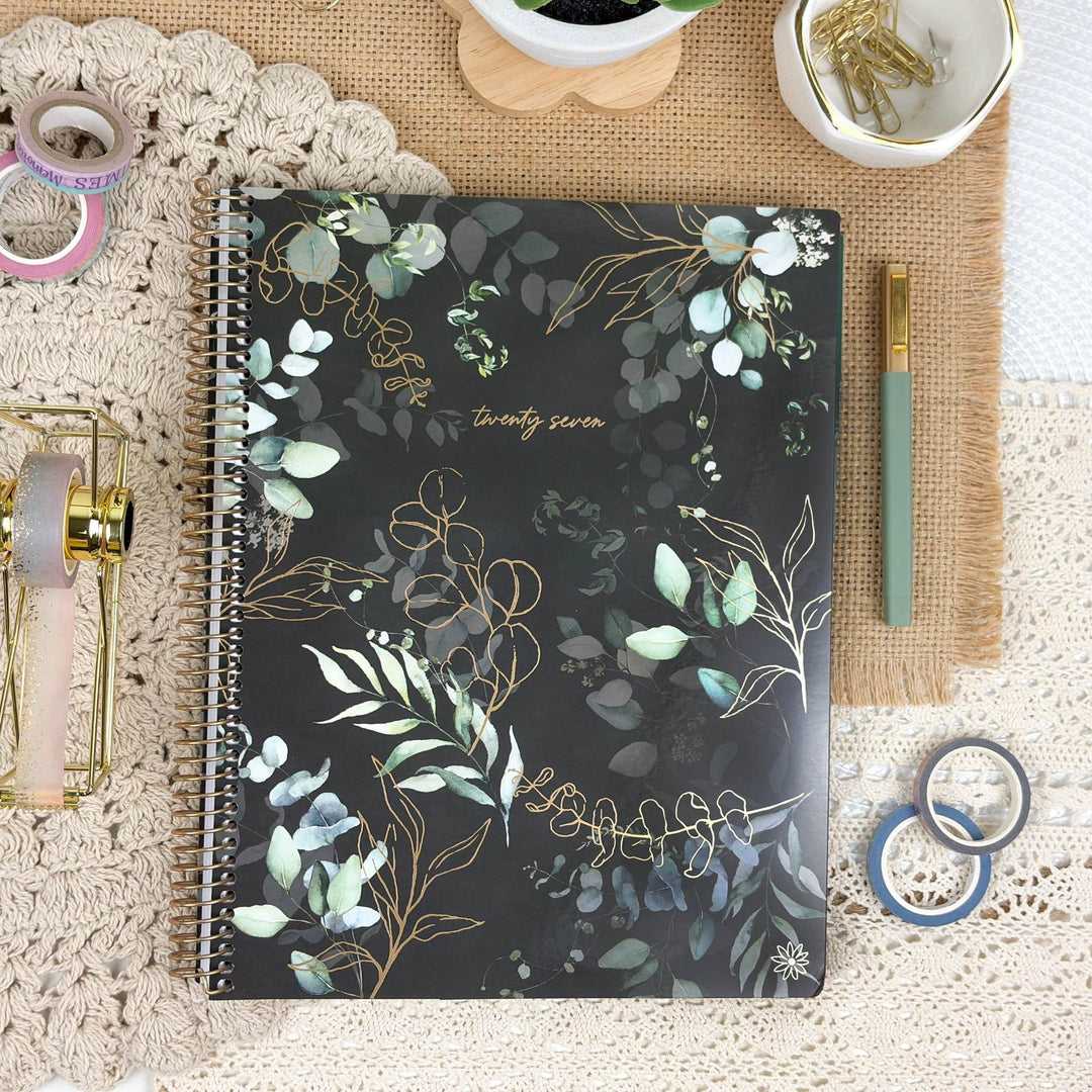 A spiral-bound notebook with a black cover decorated with gold and green botanical illustrations, styled in a flat-lay arrangement with stationery accessories on a lace tablecloth.
