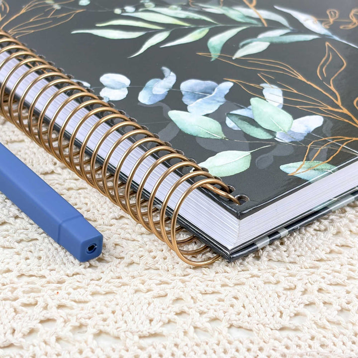 A close-up of a spiral-bound notebook with a black cover decorated with watercolor-style green and blue leaves and gold floral outlines, placed on a cream lace surface next to a blue pen.