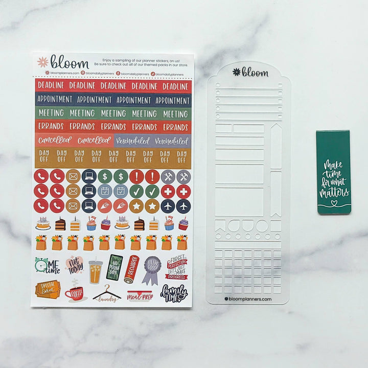 Bloom planner sticker kit with a sheet of colorful planner stickers, a white plastic date-grid planner insert, and a small green tag with white text, displayed on a white marble background.