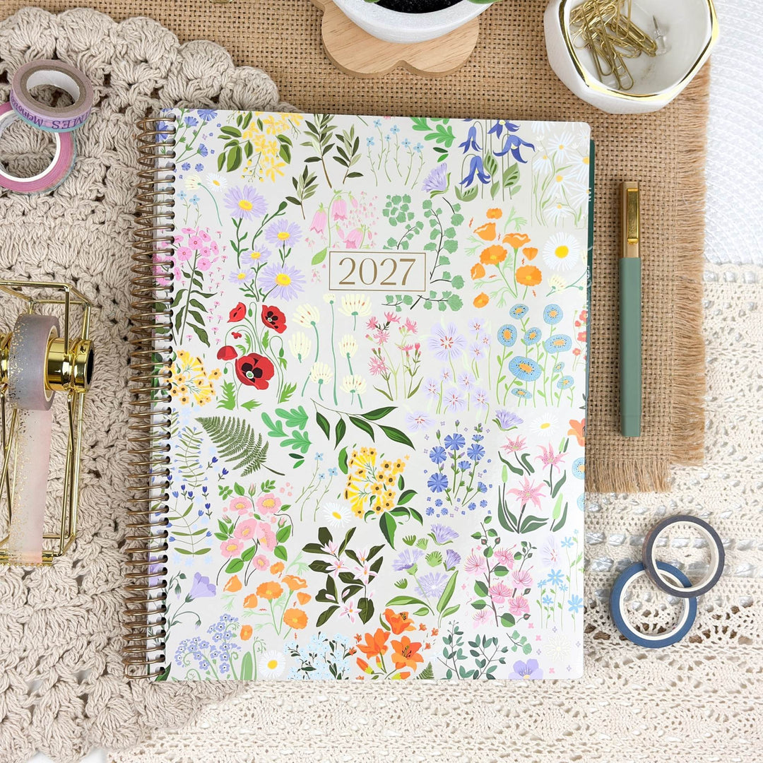 A spiral-bound 2027 floral planner lies on a beige lace surface, surrounded by stationery props including a green pen, washi tapes, a small gold container, and a bowl of gold paper clips on a burlap mat.