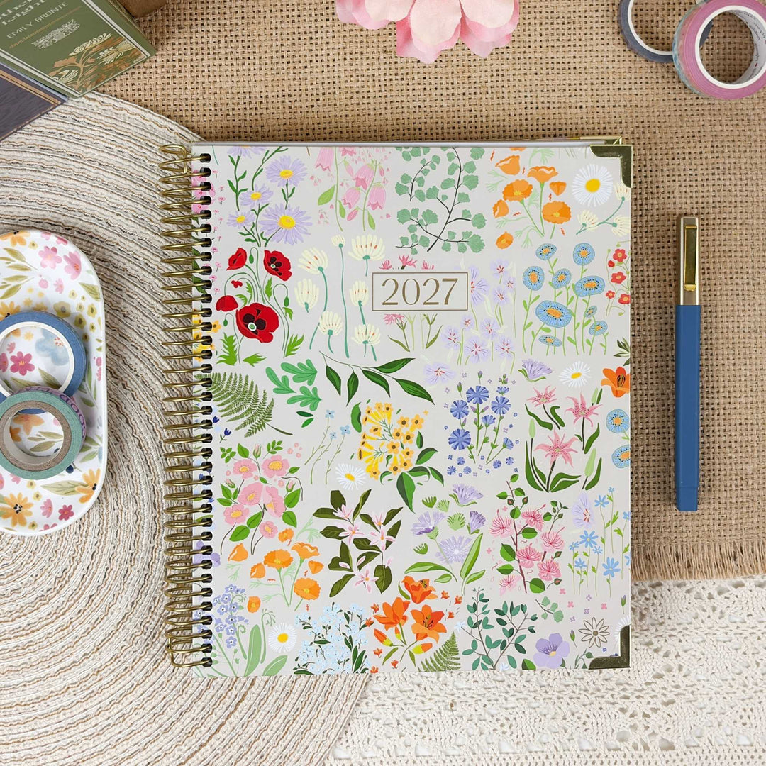 A spiral-bound 2027 floral planner lies on a burlap surface surrounded by styled desk props, including washi tape, a blue pen, a small tray, and pink flowers. The planner cover features a dense multicolor wildflower pattern on a light background.