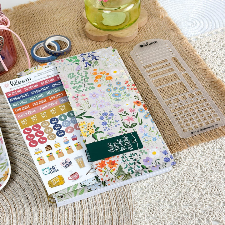 A planner sticker sheet and matching planner template set from the brand bloom, styled on a beige woven placemat with other stationery items like washi tape and a glass cup in the background.