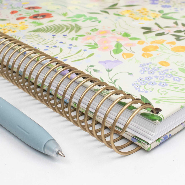 A floral-patterned spiral notebook with a gold-tone wire coil binding, shown on a white surface next to a light blue pen.