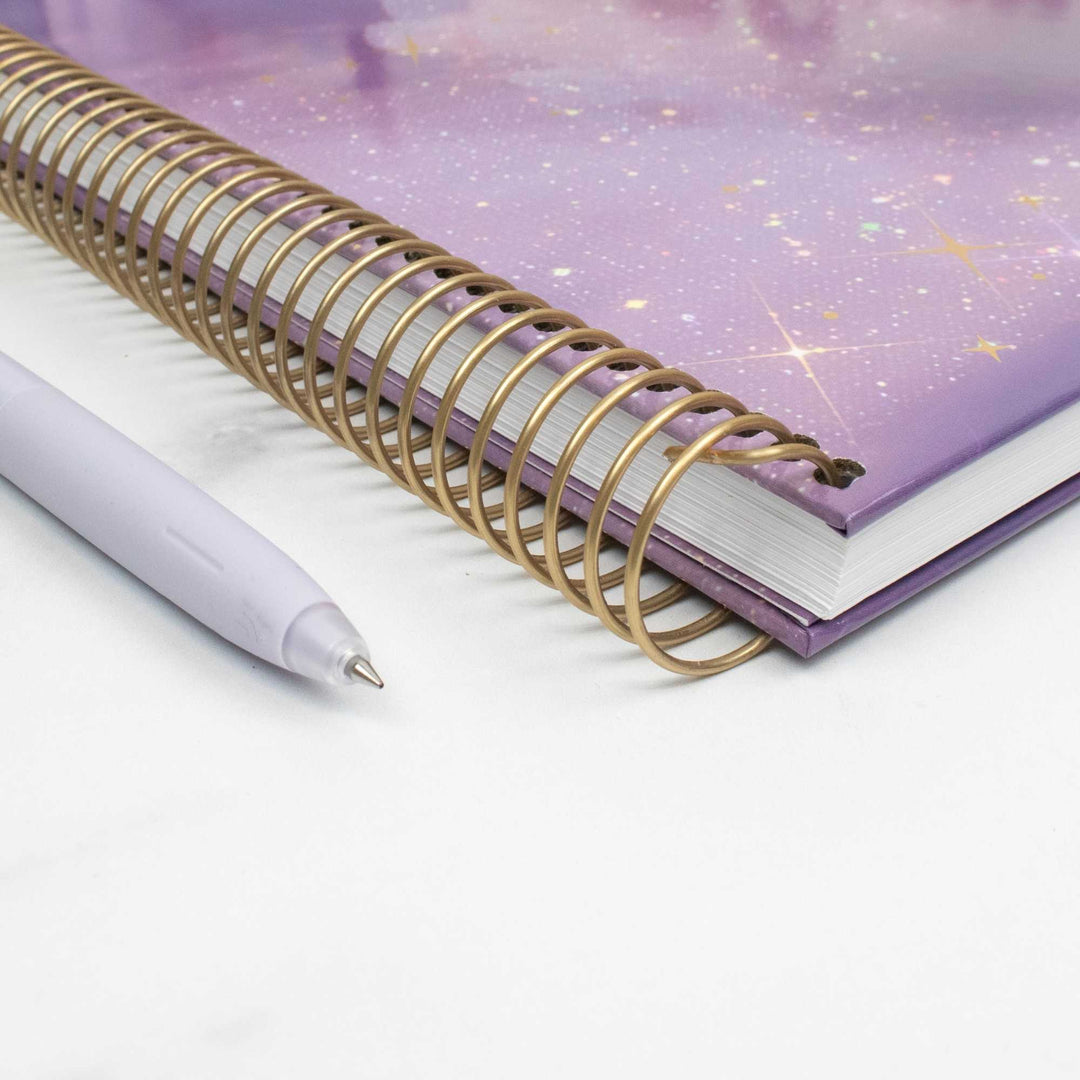 A spiral-bound notebook with a purple galaxy-patterned cover and a gold metal spiral, shown on a white surface next to a light lavender pen.