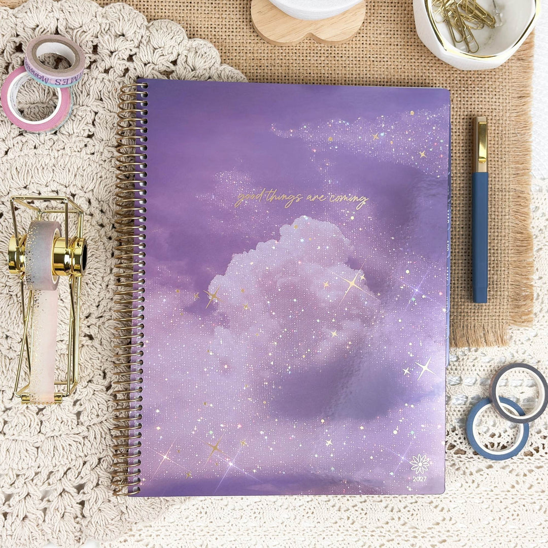A spiral-bound planner or notebook with a purple galaxy cloud cover and gold foil text reading 'good things are coming,' styled in a flat lay with stationery props on a lace cloth and burlap mat.