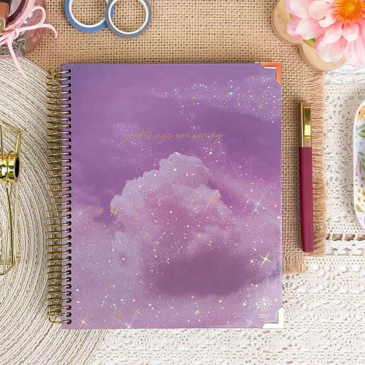 A spiral-bound planner with a purple cloud and starry sky cover, styled on a burlap mat with a pink pen, scissors, and decorative props around it.