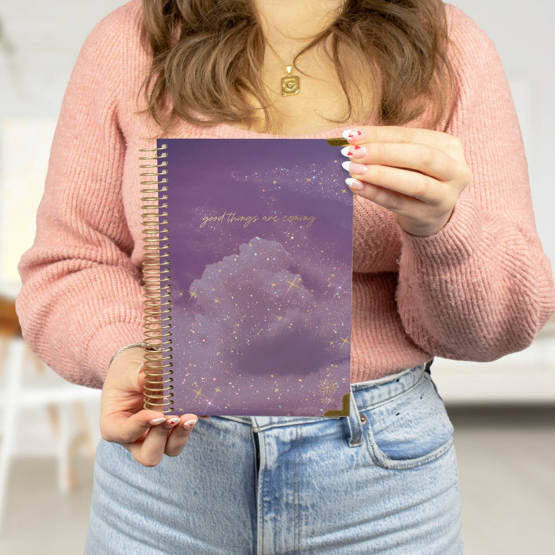 A person in a pink knit sweater holds a purple spiral notebook with a cloud and starry sky design on the cover. The notebook has gold glitter accents and the phrase 'good things are coming' in gold script.